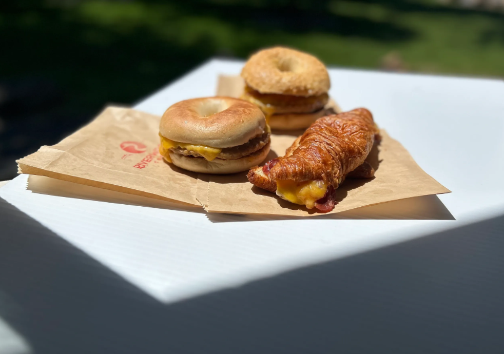 Breakfast bagel sandwiches and a bacon-and-cheddar stuffed croissant on Riverwoods-branded paper bags