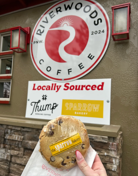 A hand holding a Sparrow Bakery chocolate chip cookie in front of the Riverwoods Coffee sign with the Locally Sourced placard featuring Thump Coffee and Sparrow Bakery logos