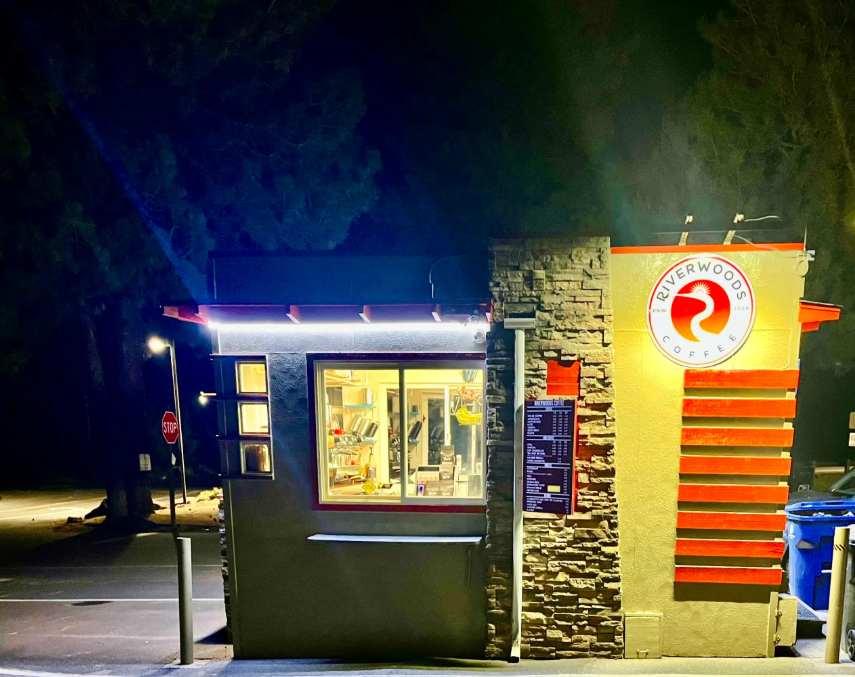 Riverwoods Coffee stand glowing at night, with warm interior light, the logo lit on the side wall, and the menu board visible to drive-thru customers