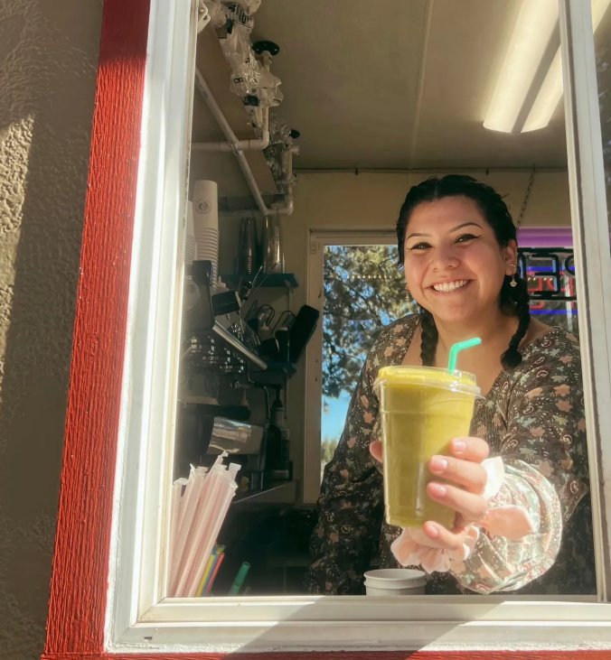 A smiling Riverwoods Coffee barista handing a fresh green smoothie out the drive-thru window