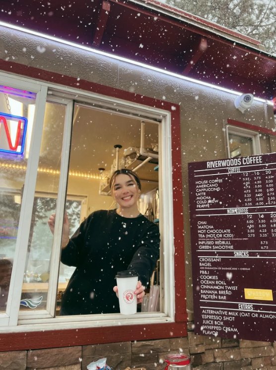 A smiling Riverwoods Coffee barista handing a cup out the window as snow falls, with the menu board alongside