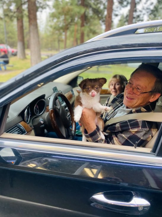 A smiling older couple in their car holding a chihuahua at the Riverwoods Coffee drive-thru, pines in the background