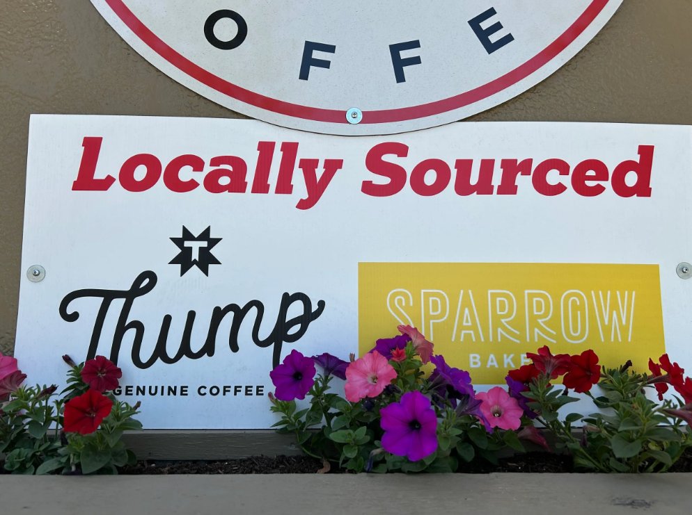 The Locally Sourced sign at Riverwoods Coffee featuring the Thump Coffee and Sparrow Bakery logos, framed by colorful petunias