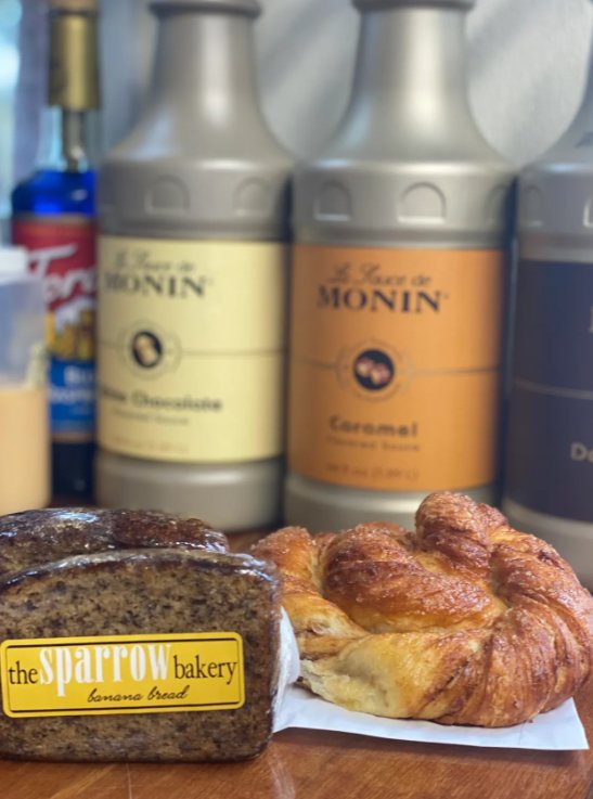 Sparrow Bakery banana bread and a croissant beside Monin syrup bottles inside Riverwoods Coffee