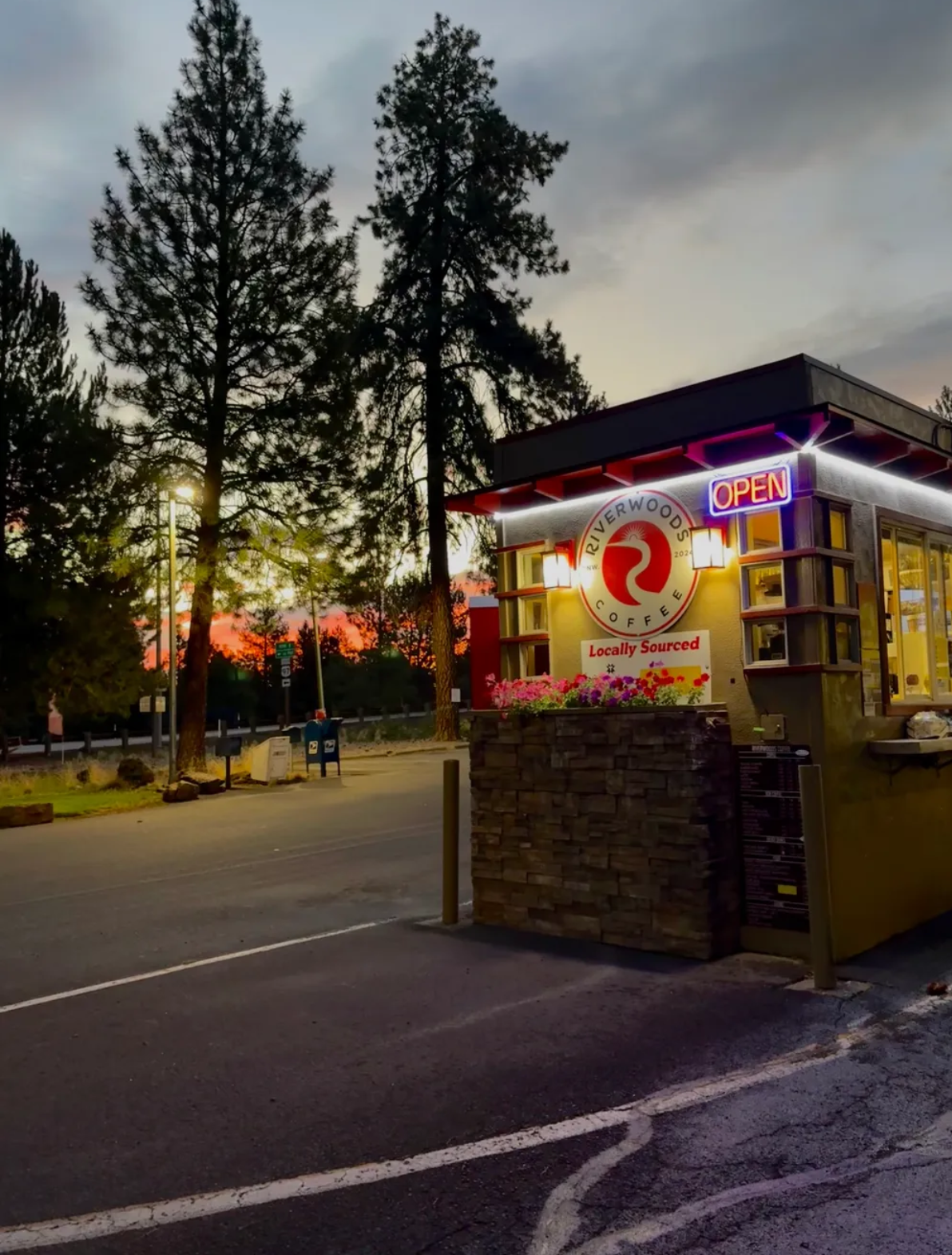 Riverwoods Coffee stand glowing under tall pines at sunset with neon OPEN sign